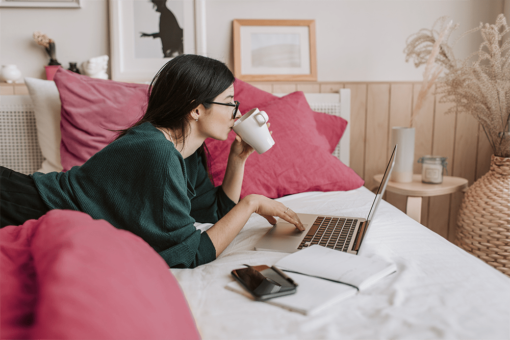 Žena ležiaca na posteli. Číta blog na notebooku a pije pri tom čaj.