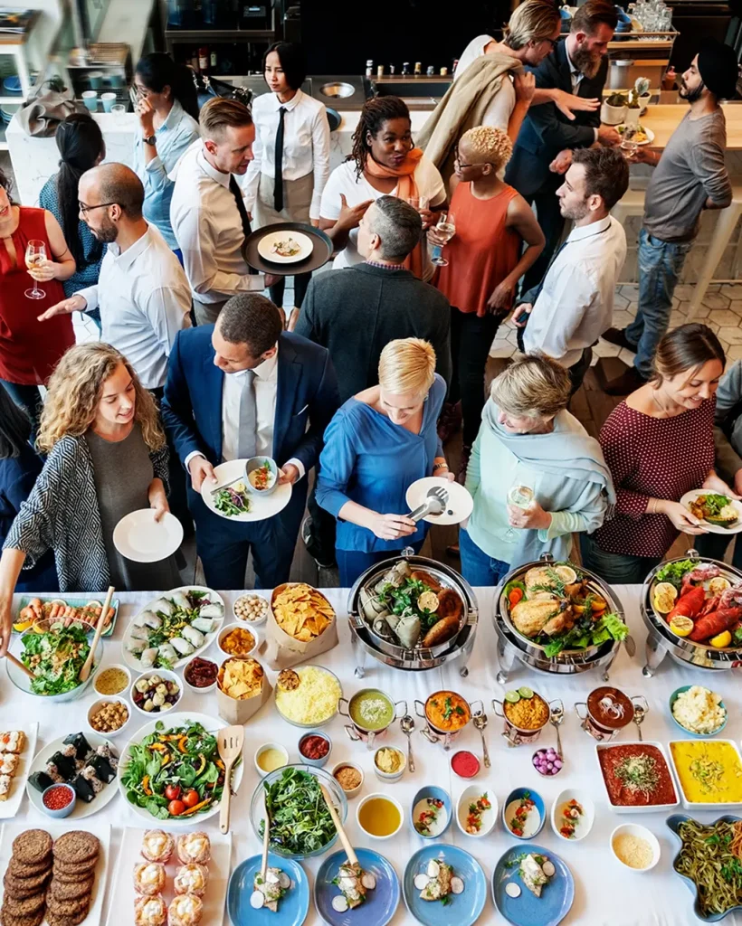 Švédske stoly a skupina ľudí s taniermi a pohármi. Medzi hosťami chodia častníci. Ľudia debatujú.