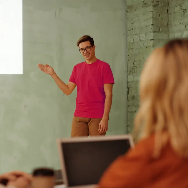 Muž ukazujúci na stenu, na ktorej je prezentácia z projektoru. V popredí sedí žena robiaca si poznámky na notebooku.