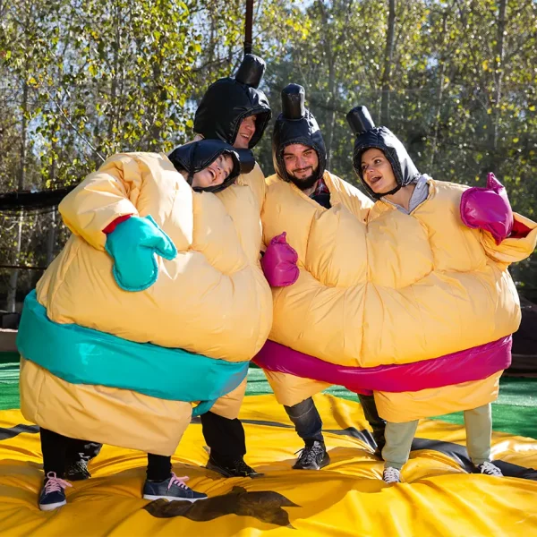 teambuilding Štyria ľudia v žltých nafukovacích sumo oblekoch. V jednom nafukovacom obleku sú dvaja ľudia. Stoja na mäkkej podložke.