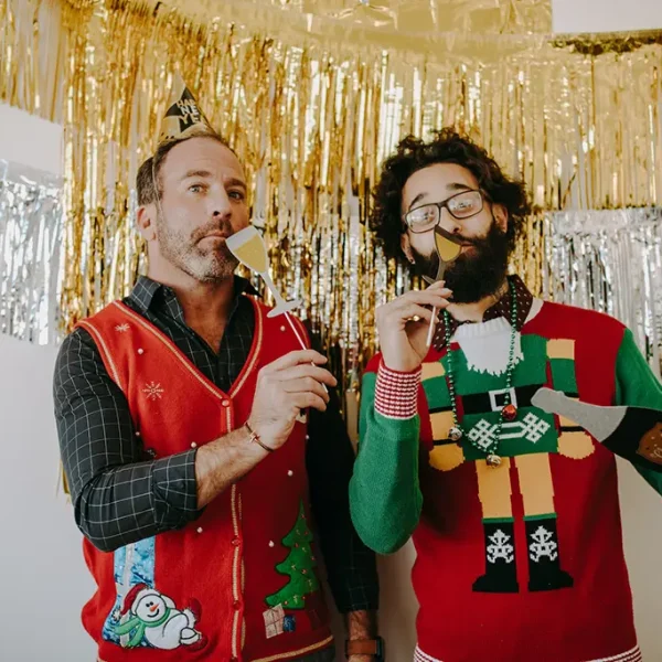 Dvaja muži vo vianočných svetroch s rekvizitami na fotenie. V pozadí je strieborno-zlatá výzdoba